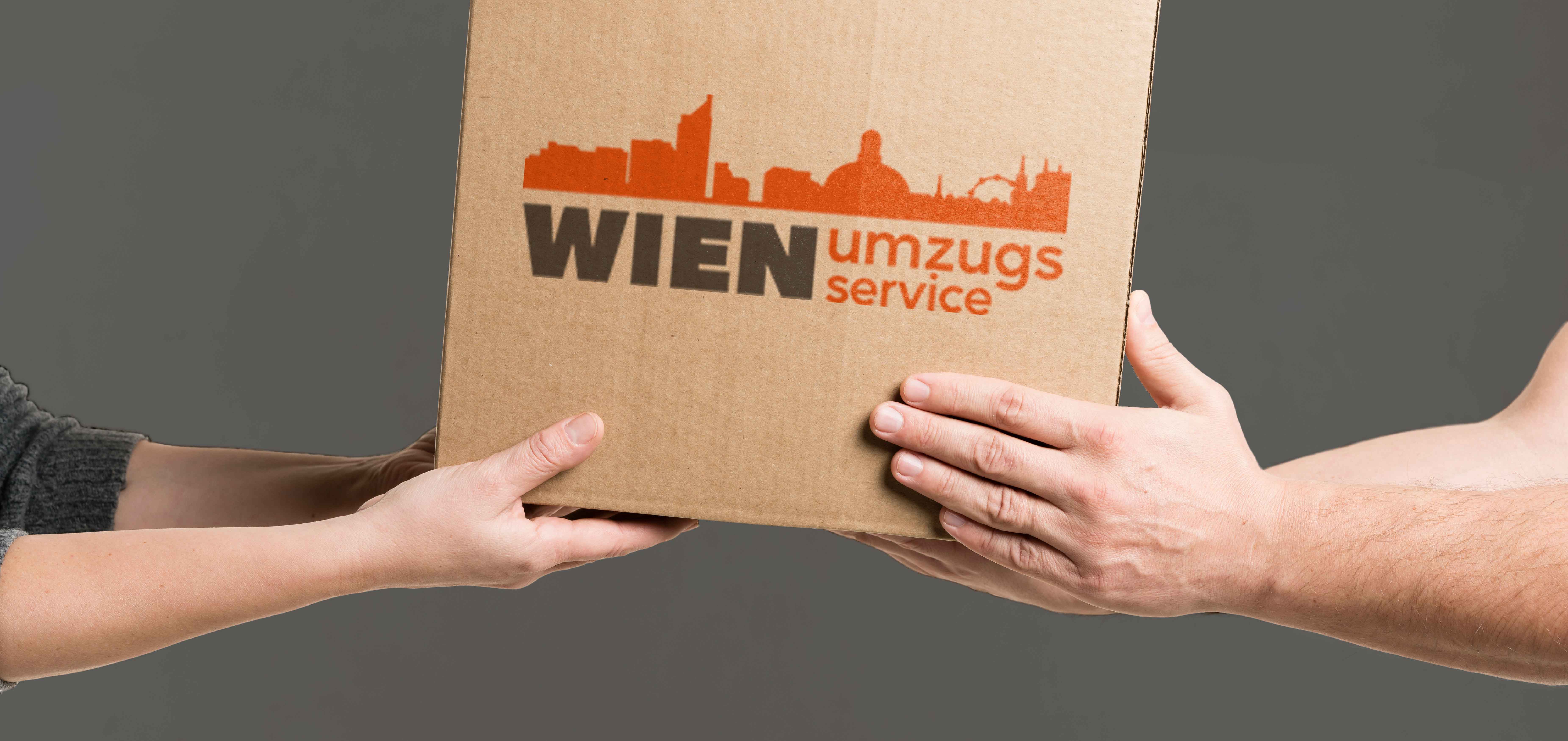 Kisten mit der Aufschrift 'Wien Umzugsservice' befinden sich an der Wand übereinander. An der Wand hängt ein Logo der Firma 'Wien Umzugsservice' in Orange und Grau